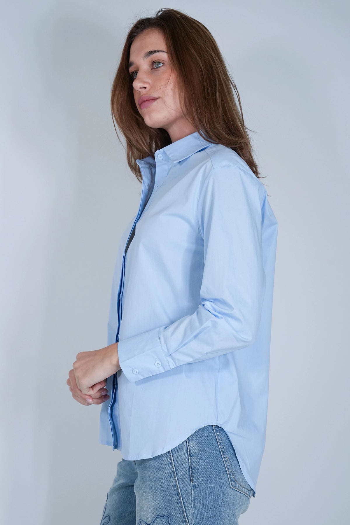 A woman wearing a light blue long-sleeve button-up shirt and light-wash blue jeans, shown in a side-profile view against a plain light gray studio background.