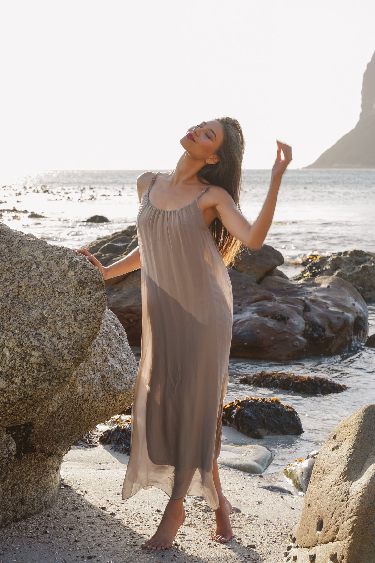 Woman in a flowing dress standing on a rocky beach with ocean and sky in the background