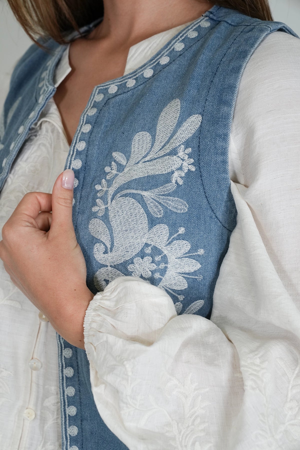 A close-up of a person wearing a blue denim vest with white floral embroidery over a white long-sleeve blouse.