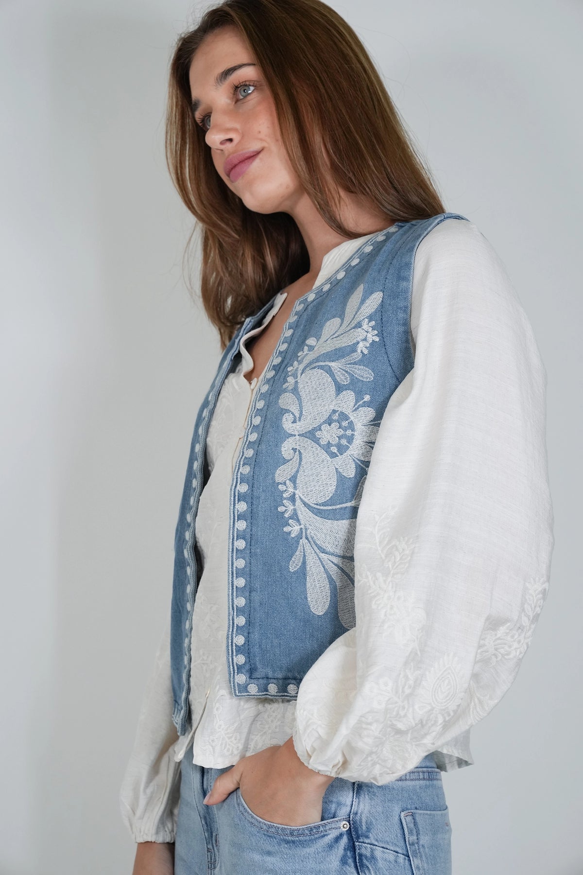 A woman wearing a light blue denim vest with white floral embroidery over a white long-sleeve blouse and light blue jeans, photographed against a plain light gray background.