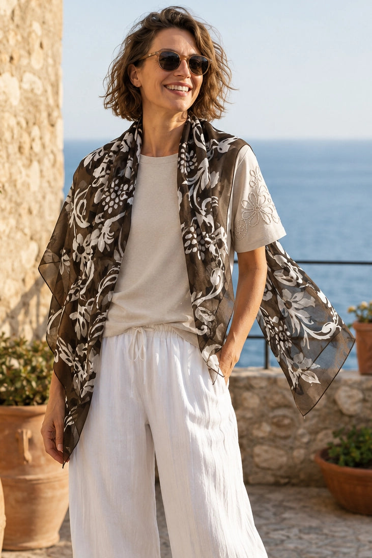 A woman wearing a sheer brown and white floral print shawl over a cream t-shirt and white wide-leg pants, standing outdoors with the sea and stone walls in the background.