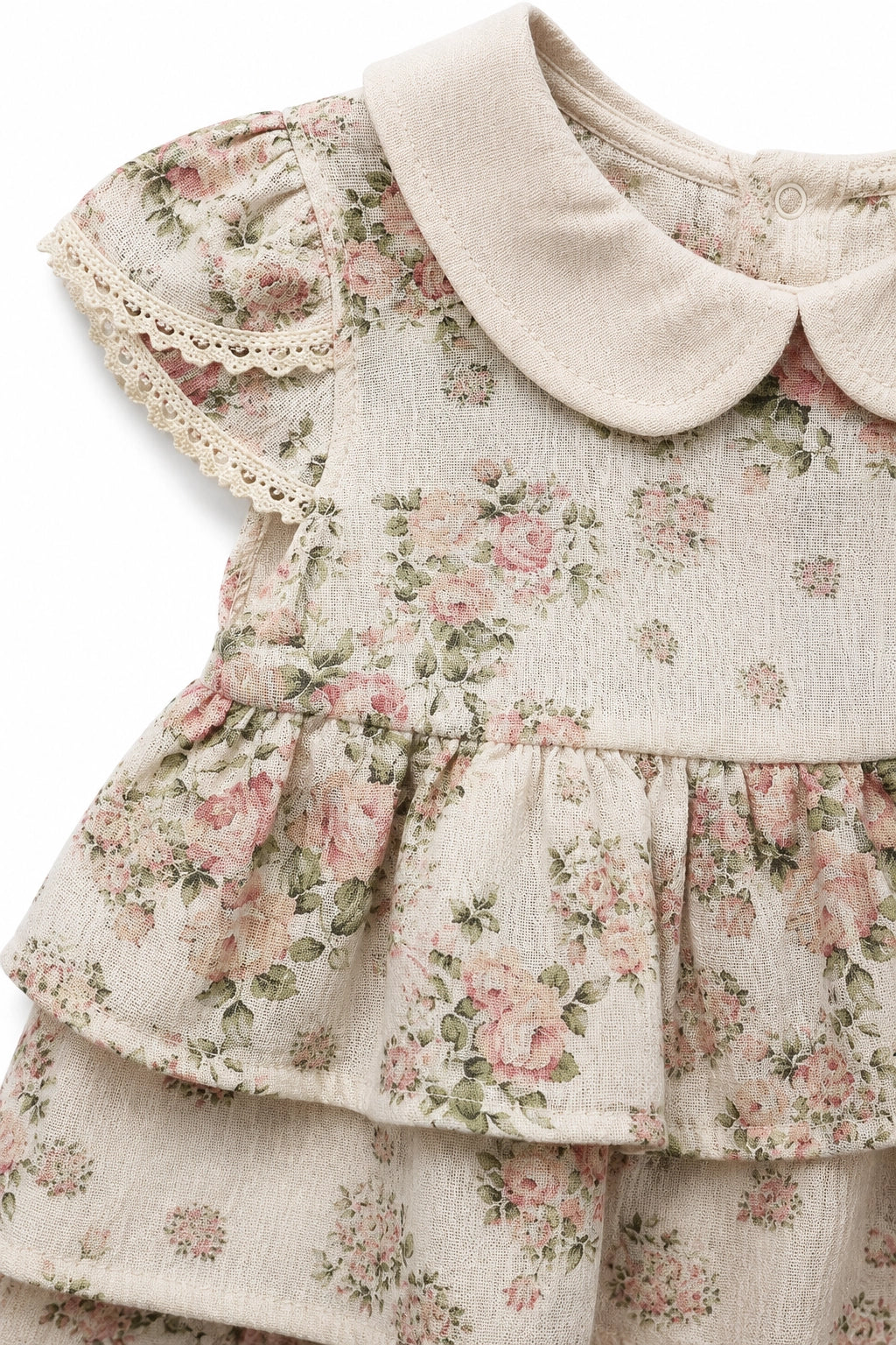 A close-up of a baby girl's floral dress featuring a cream-colored Peter Pan collar, flutter sleeves with lace trim, and a tiered ruffle bodice with a pink and green rose print on a textured cream fabric.