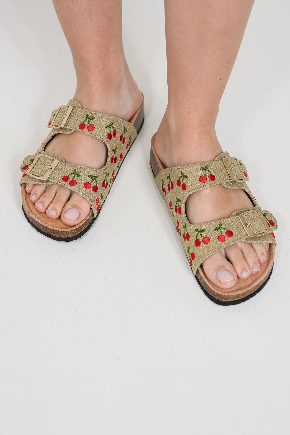 A pair of beige double-strap slide sandals with an embroidered cherry pattern, shown on a person's feet against a plain white background.