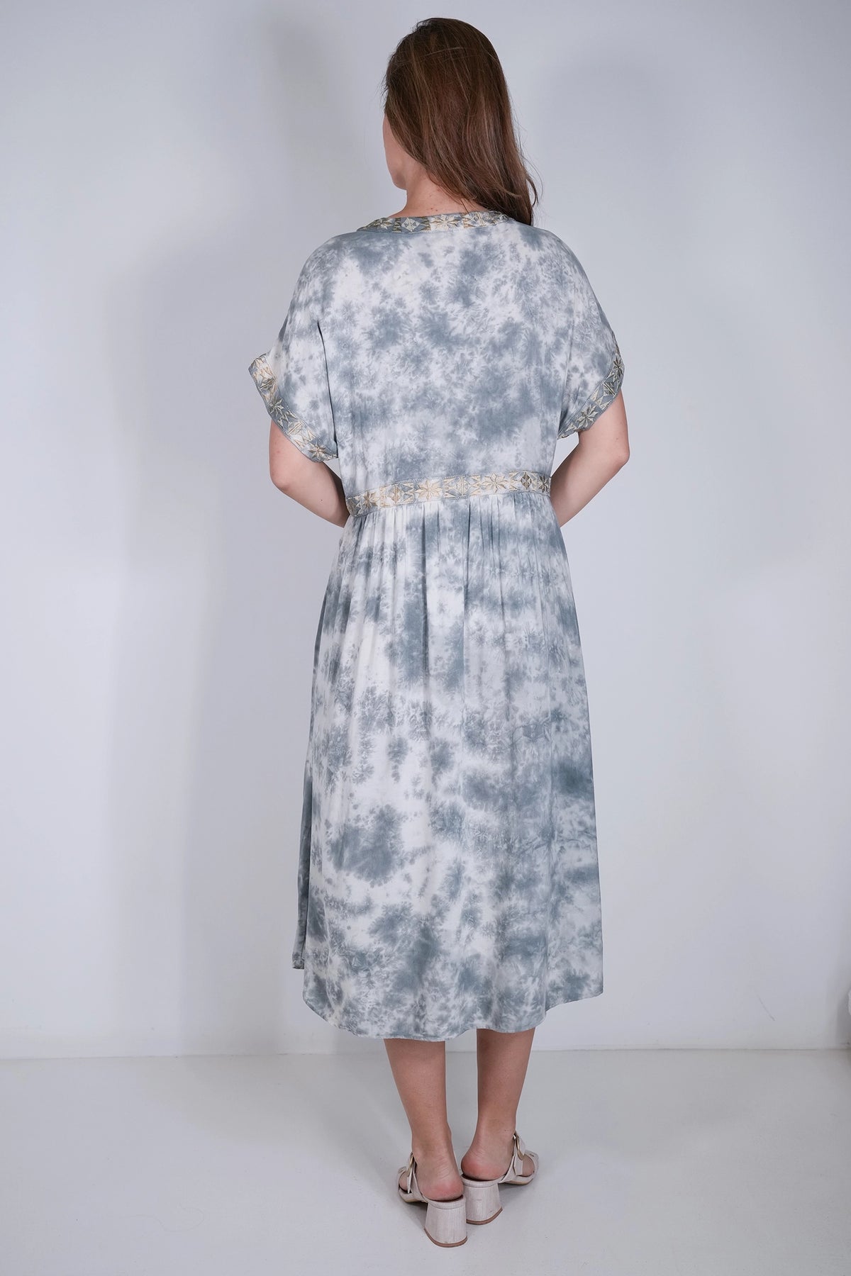 A woman viewed from behind wearing a loose, knee-length dress with a blue and white tie-dye pattern and a light beige waistband, standing against a plain light gray studio background.