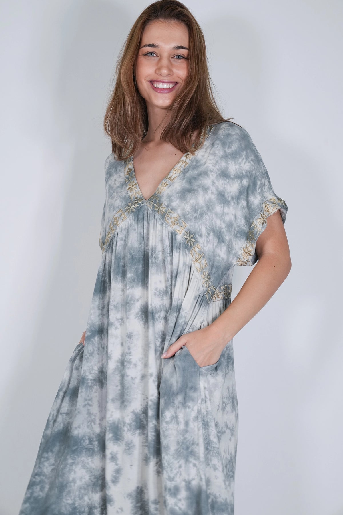 A woman modeling a loose, short-sleeve maxi dress with a blue and white tie-dye pattern and gold embroidered trim along the neckline and sleeve cuffs, set against a plain light gray studio background.