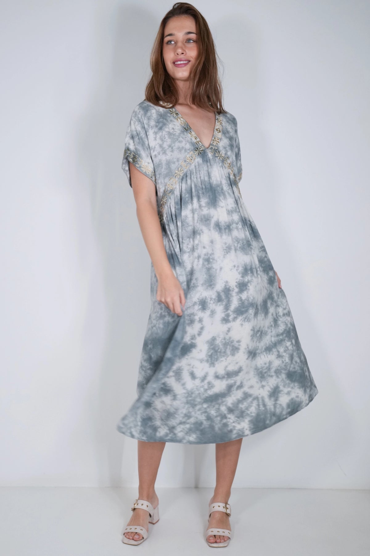 A woman modeling a short-sleeve midi dress with a blue and white tie-dye pattern and gold trim along the V-neckline, set against a plain white studio background.