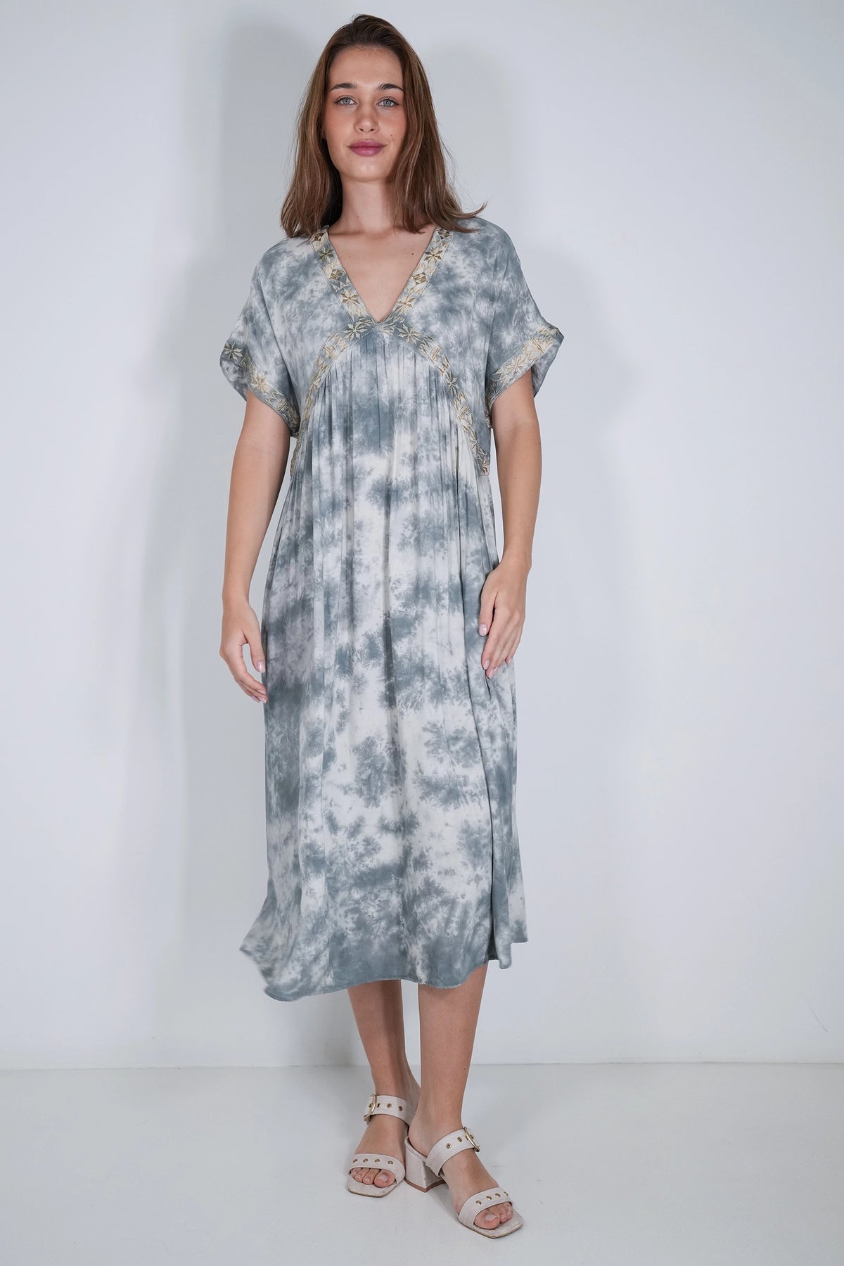A woman modeling a short-sleeve midi dress with a blue and white tie-dye pattern, featuring a V-neckline with gold embroidery details, set against a plain light gray studio background.