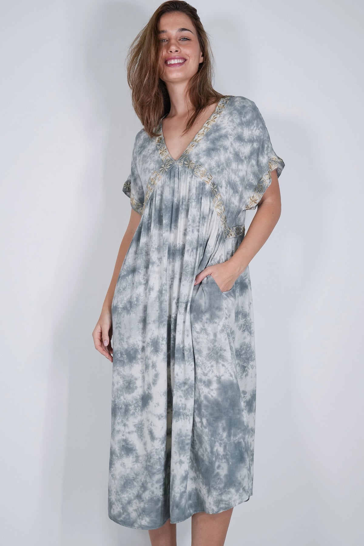 A woman modeling a loose, knee-length kaftan dress with a gray and white tie-dye pattern and gold embroidered trim along the neckline and sleeve edges, set against a plain light gray studio background.