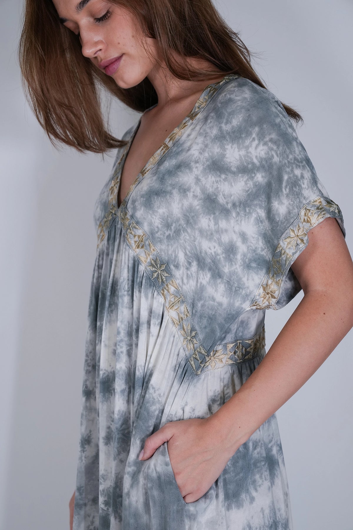 A woman wearing a gray and white tie-dye kaftan dress with gold embroidered trim, shown in a close-up side profile against a light gray background.
