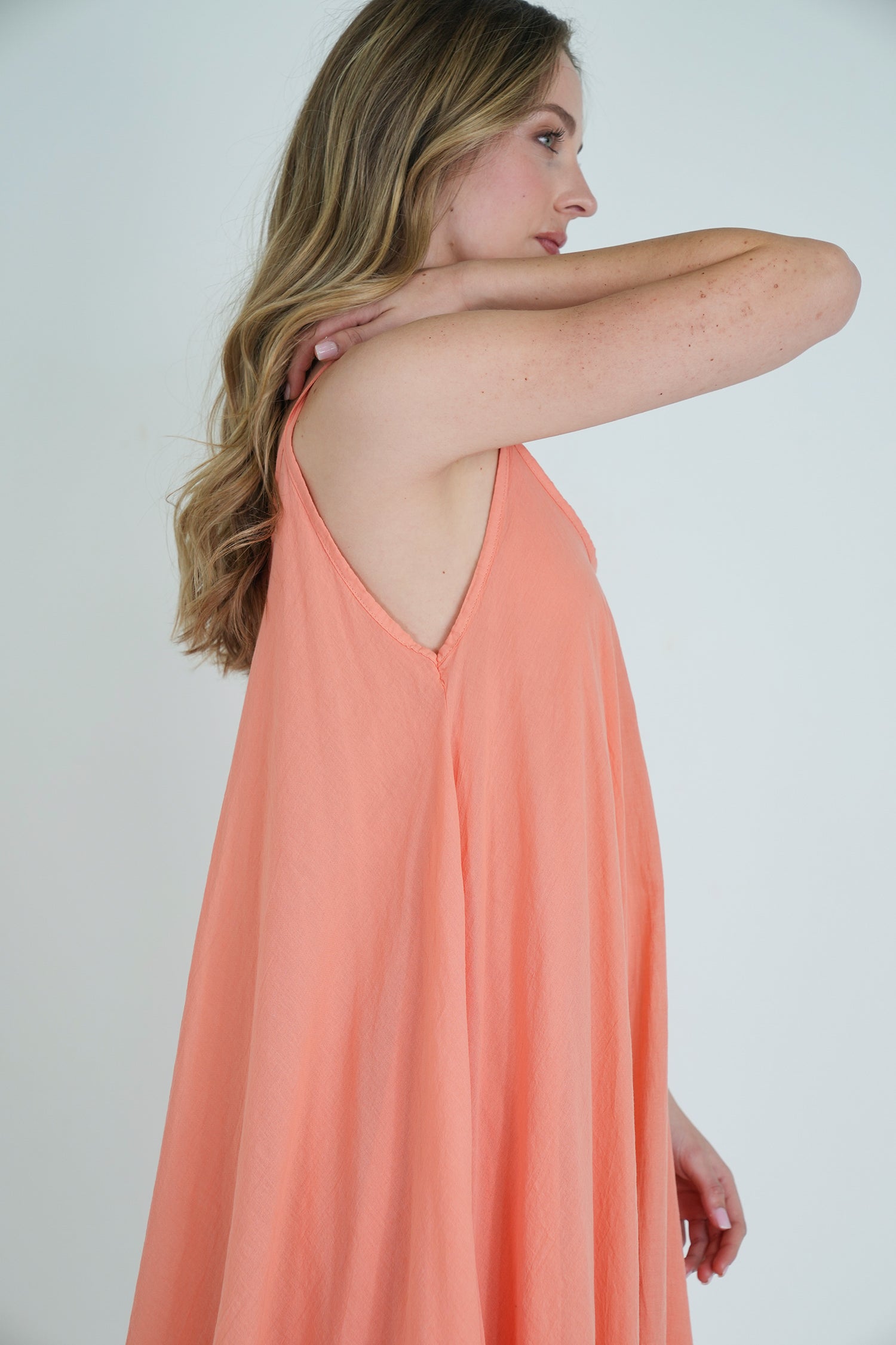 Side view of model wearing a sleeveless coral flowy dress with a relaxed fit and soft fabric texture, posing against white background.