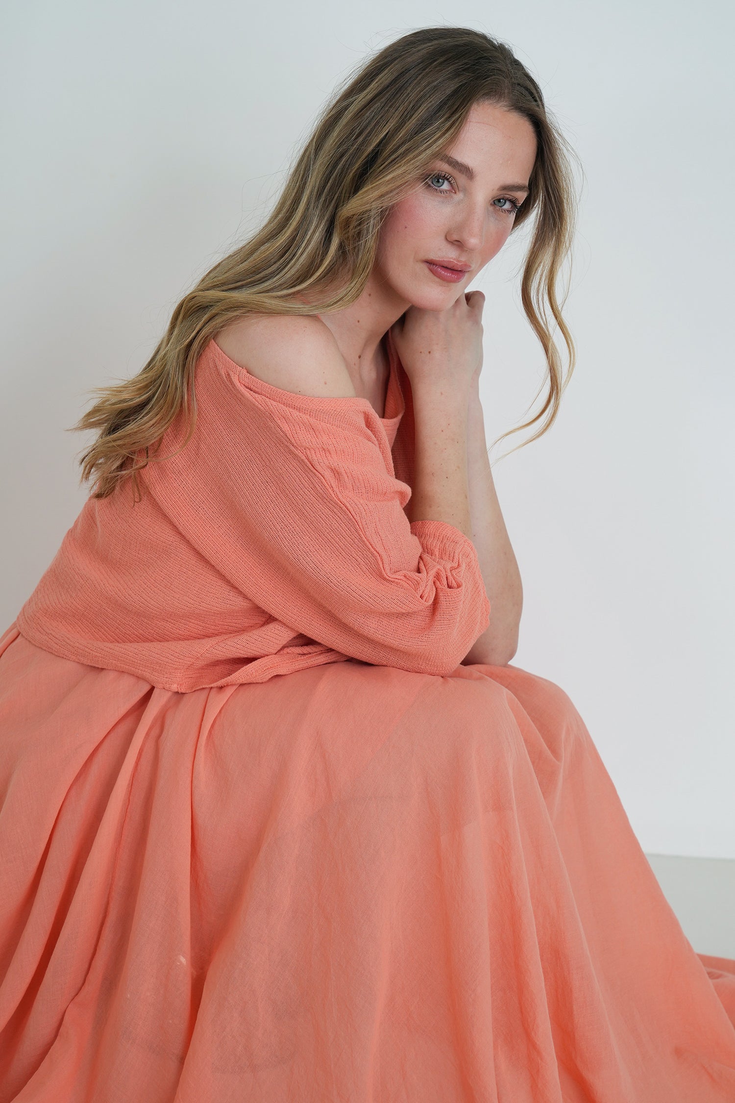 Model wearing coral flowy dress with long sleeves, sitting with legs folded, smiling softly indoors.