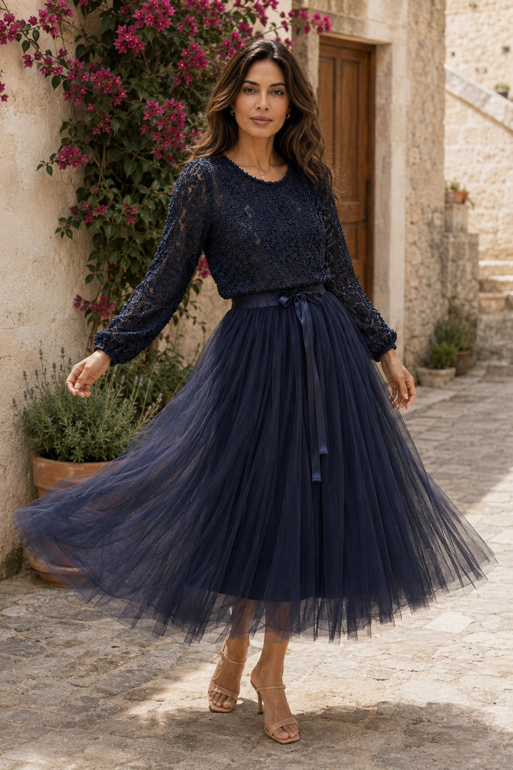 A woman wearing a navy blue lace long-sleeve top and matching navy tulle midi skirt with a tie waist, standing in a stone alleyway with flowers and a rustic building in the background.