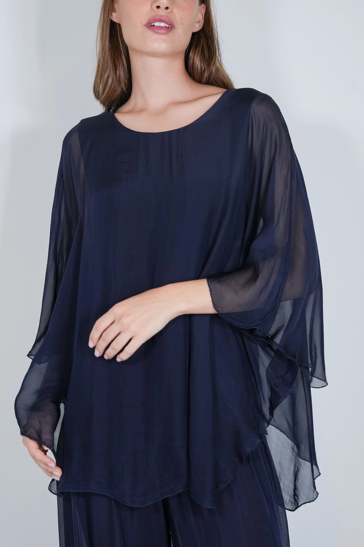A woman wearing a navy blue sheer long-sleeve top with a wide scoop neckline and layered flutter sleeves, paired with matching navy pants, against a light gray studio background.