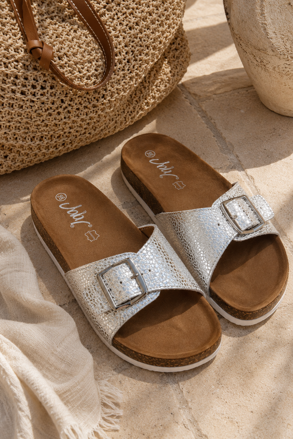 A pair of women's slide sandals with textured silver straps and square buckles, brown footbeds, and cork soles, styled on a stone surface with a woven bag and cream fabric.