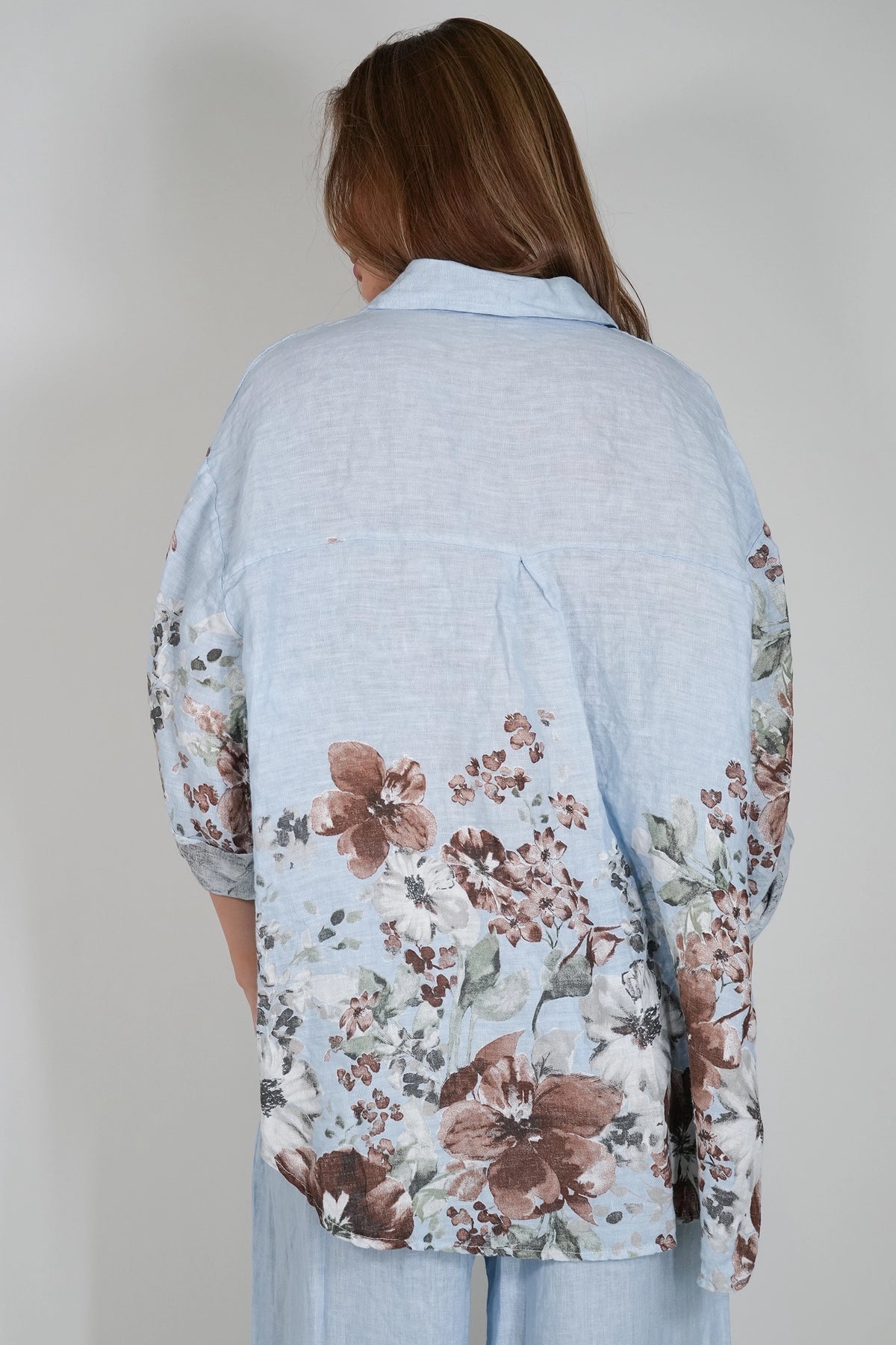 A woman viewed from behind wearing a light blue linen button-up shirt with a brown and white floral print on the sleeves and lower back, paired with light blue pants against a neutral gray background.