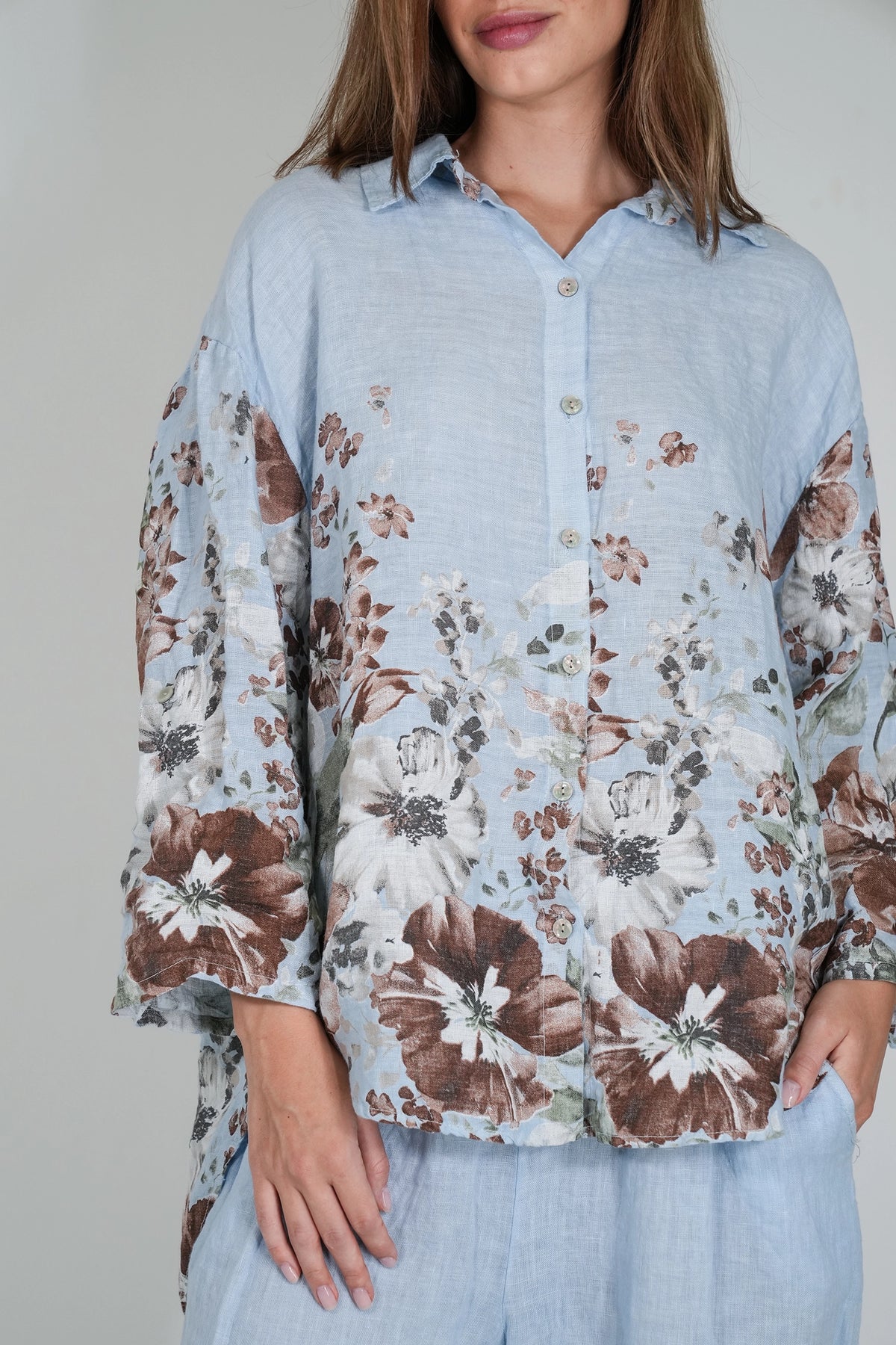 A woman wearing a light blue button-up shirt with a brown and white floral print, paired with matching light blue pants, against a plain light gray background.