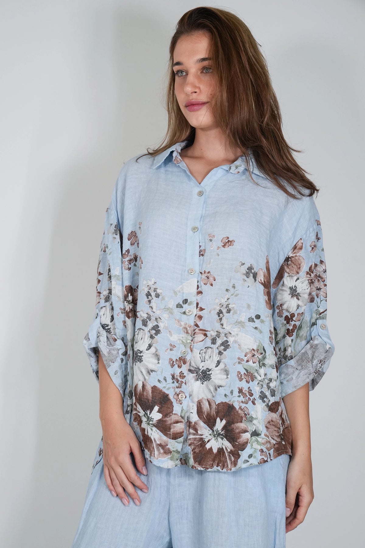A woman wearing a light blue button-up shirt with a brown and white floral print, paired with matching light blue trousers, photographed against a plain light gray background.