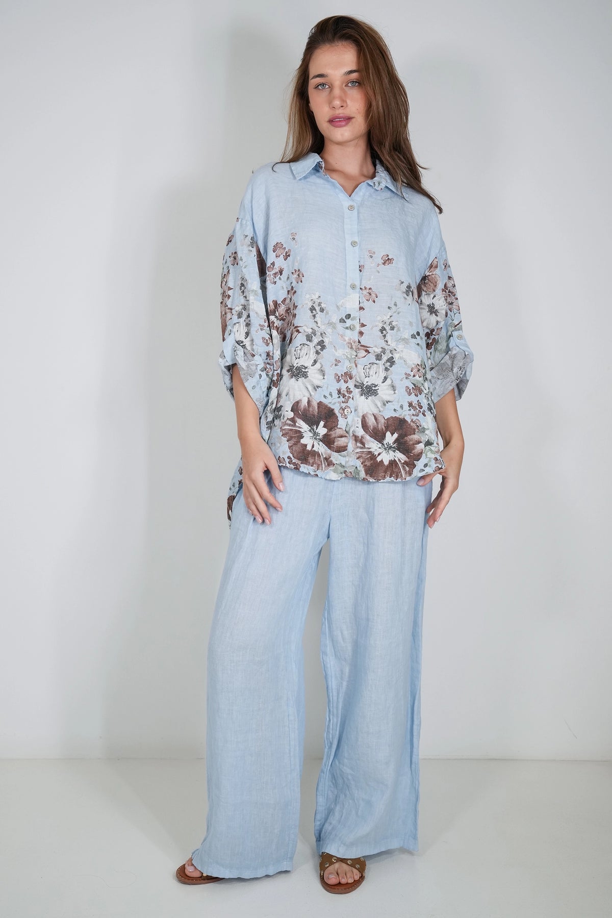 A woman modeling a light blue button-up shirt with a brown and white floral print, paired with matching wide-leg light blue pants, against a plain light gray studio background.