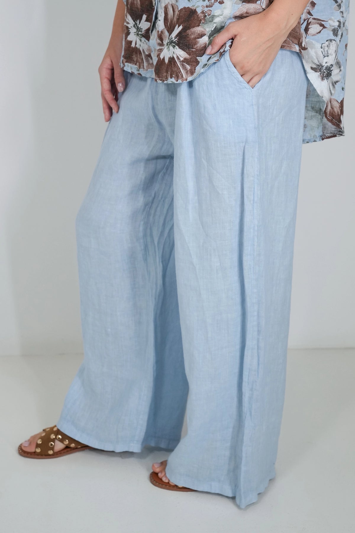 A person wearing light blue wide-leg linen pants, paired with a brown and white floral top and brown slide sandals, shown from the waist down against a plain white studio background.