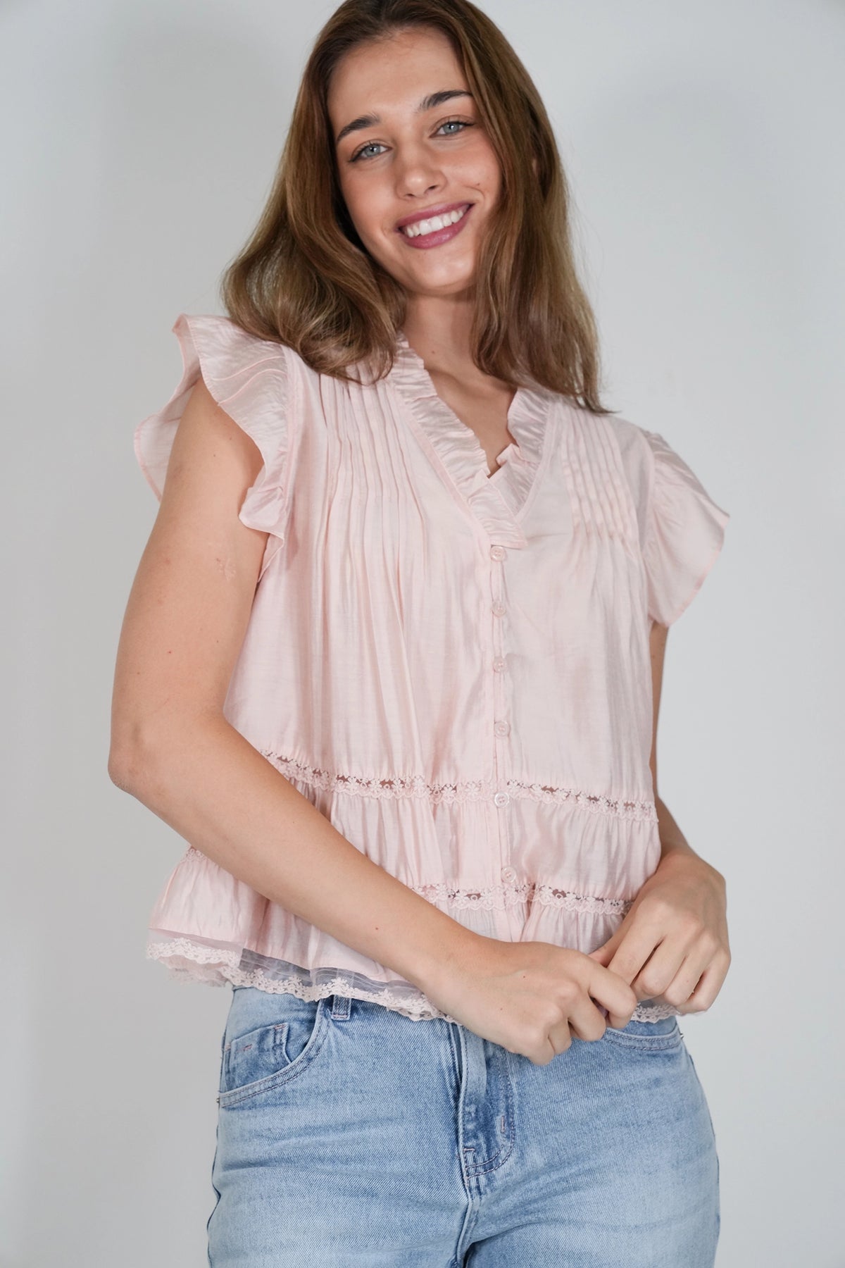 A woman wearing a light pink sleeveless blouse with ruffle trim and a tiered peplum hem, paired with light blue jeans, against a light gray studio background.