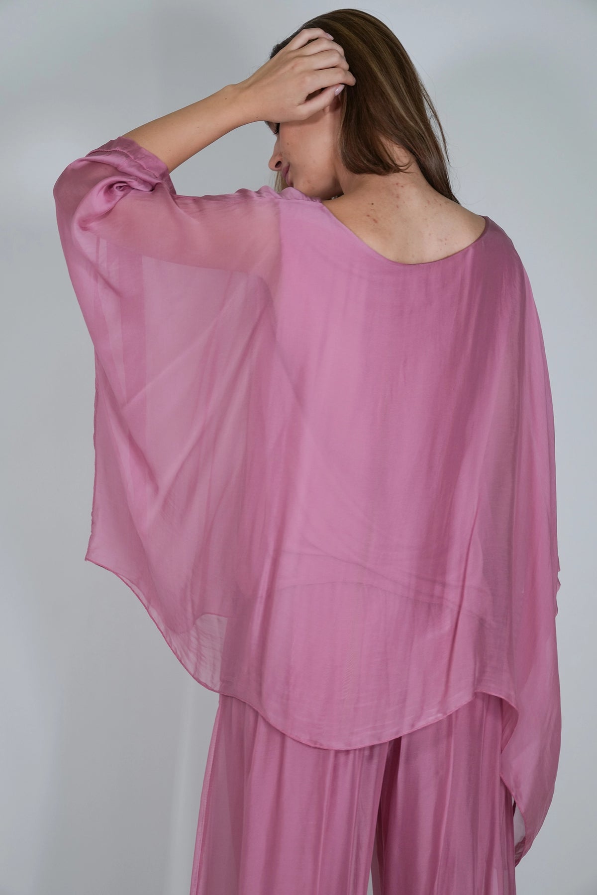 A woman wearing a loose, flowing dusty pink sheer top and matching pants, photographed from the back against a plain light gray studio background.