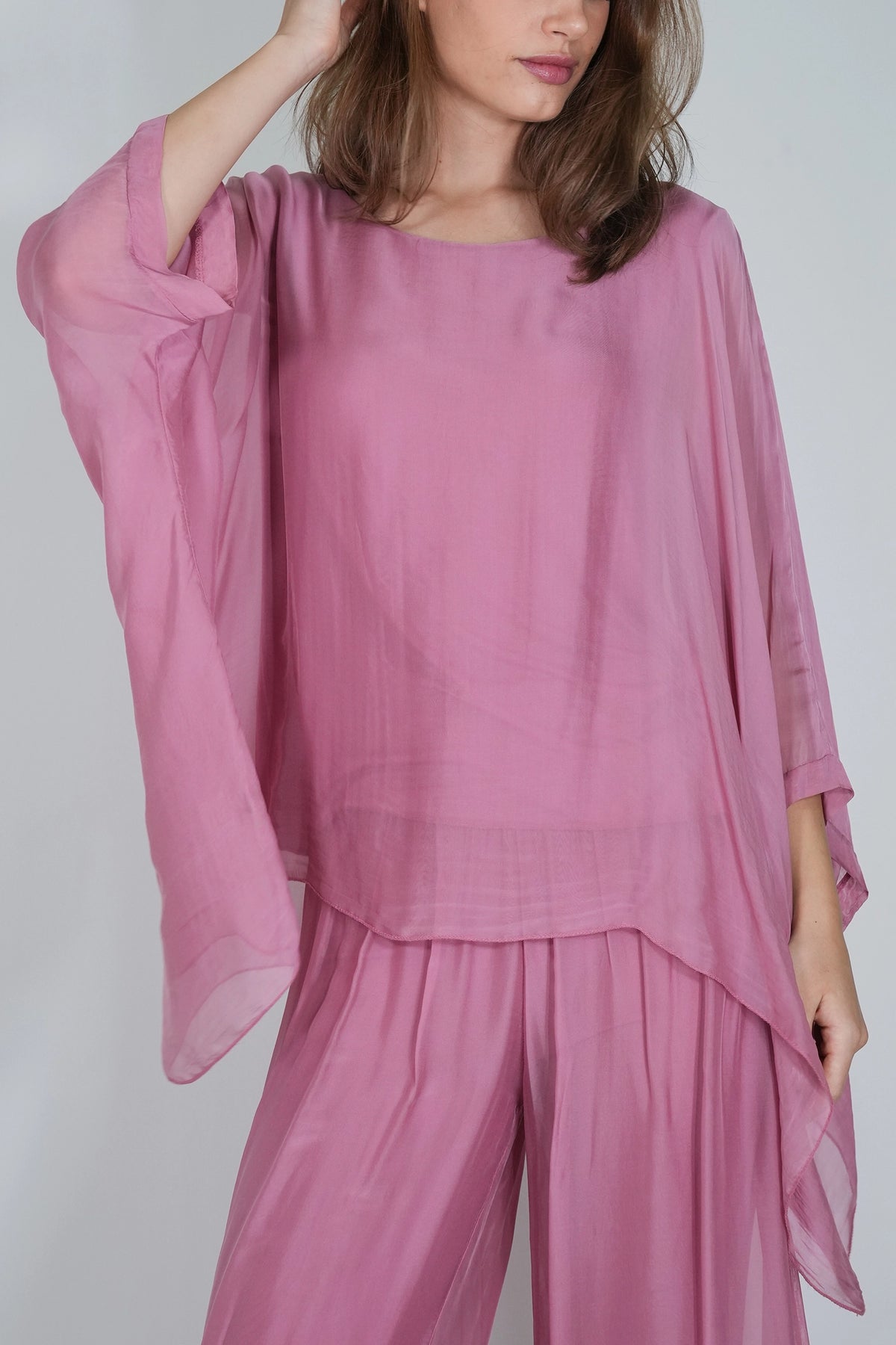 A woman wearing a loose, flowing dusty pink two-piece outfit with a sheer long-sleeve top and matching pleated pants, photographed against a plain light gray background.