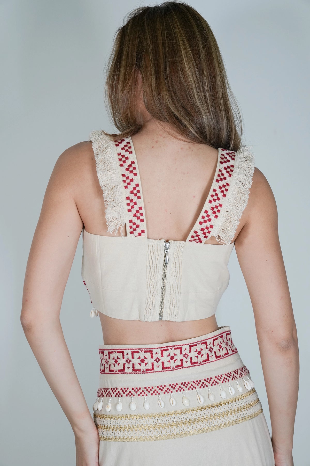 A woman shown from the back wearing a cream-colored embroidered crop top and matching skirt with red and white geometric patterns and fringe details.