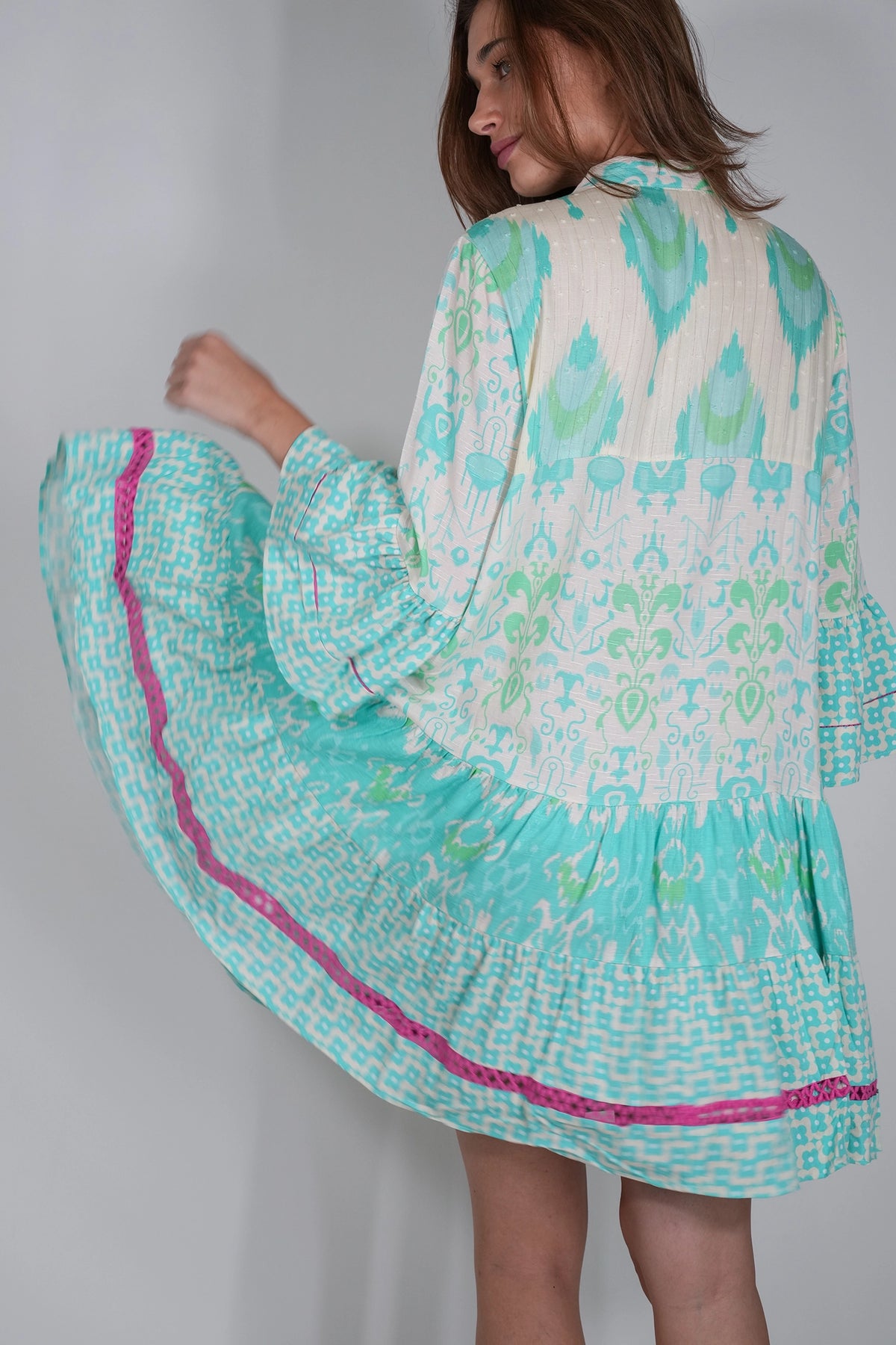 A woman wearing a long-sleeve tiered dress with a white base and mint green ikat-style patterns, featuring a bright pink trim along the skirt, shown from a side-back angle against a plain light gray wall.