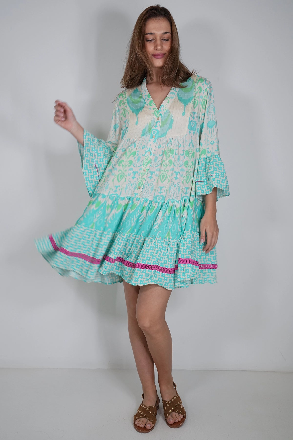 A woman modeling a short, tiered dress with a green and white tropical leaf pattern, featuring a V-neckline, three-quarter bell sleeves, and a bright pink patterned hem trim, set against a plain light gray studio background.