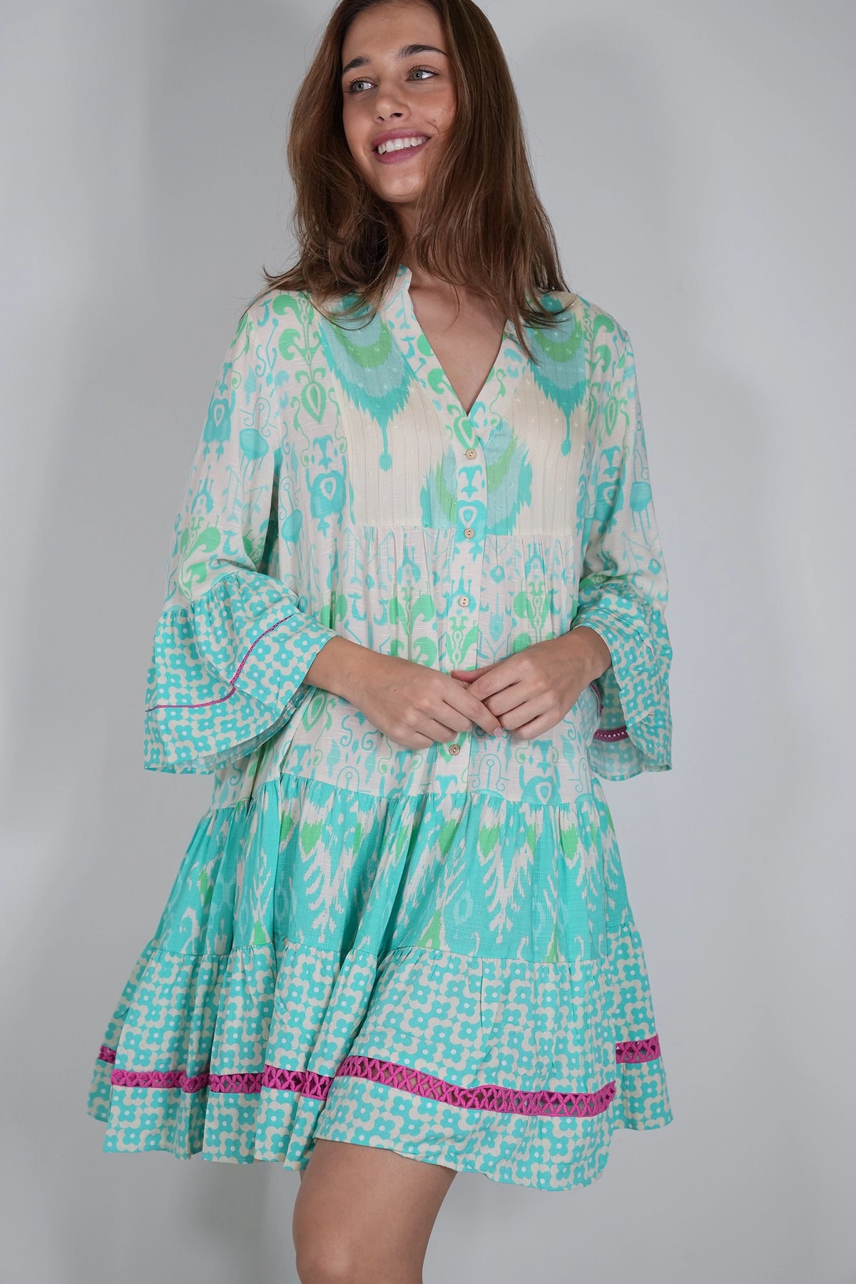 A woman modeling a short tiered boho-style dress with a turquoise, mint green, and cream ikat pattern, featuring pink patterned trim at the hem and bell sleeves.