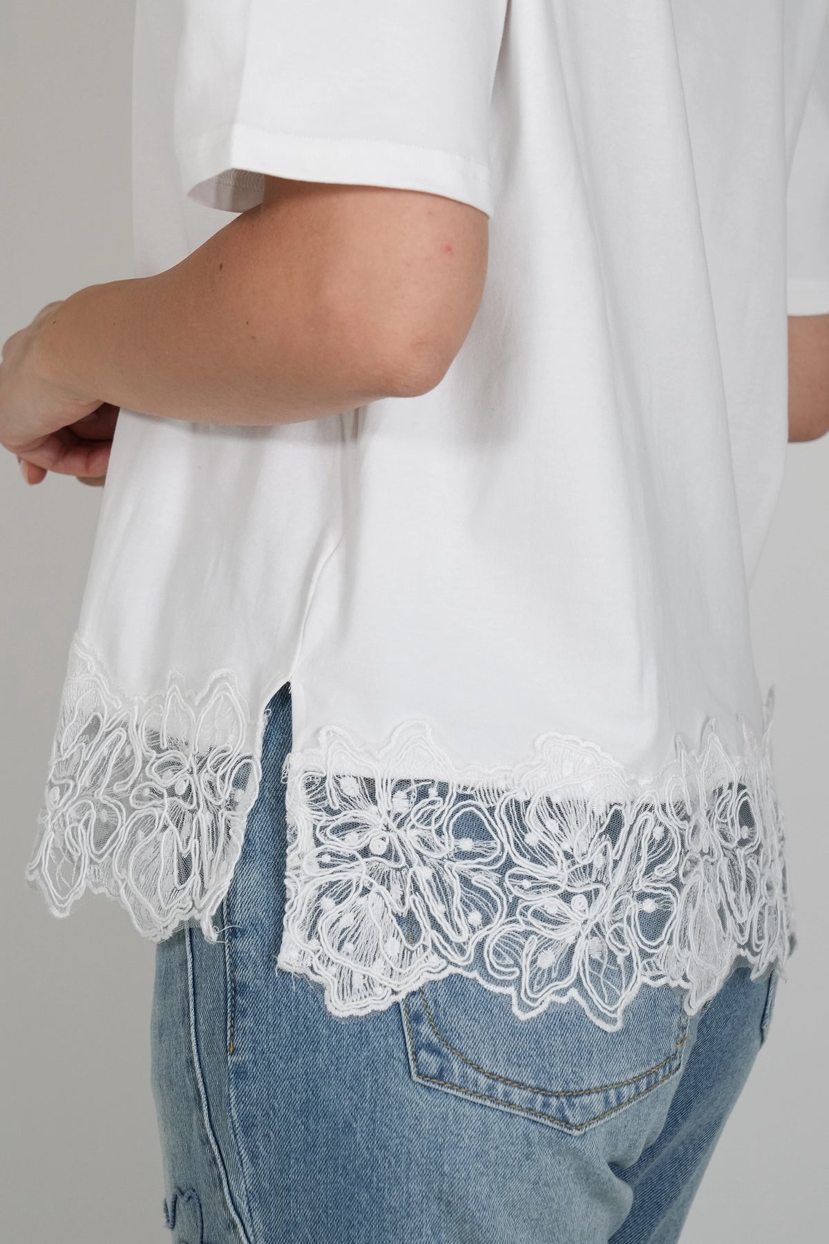 White short-sleeve blouse with a floral lace hem, shown on a model wearing blue jeans against a light gray background.