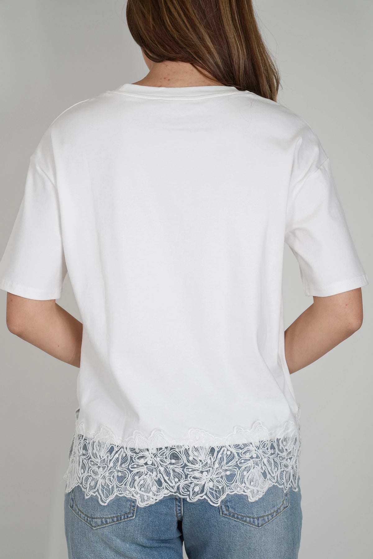 A woman viewed from the back wearing a white short-sleeve T-shirt with a decorative white lace hem, paired with blue jeans, against a plain light gray studio background.