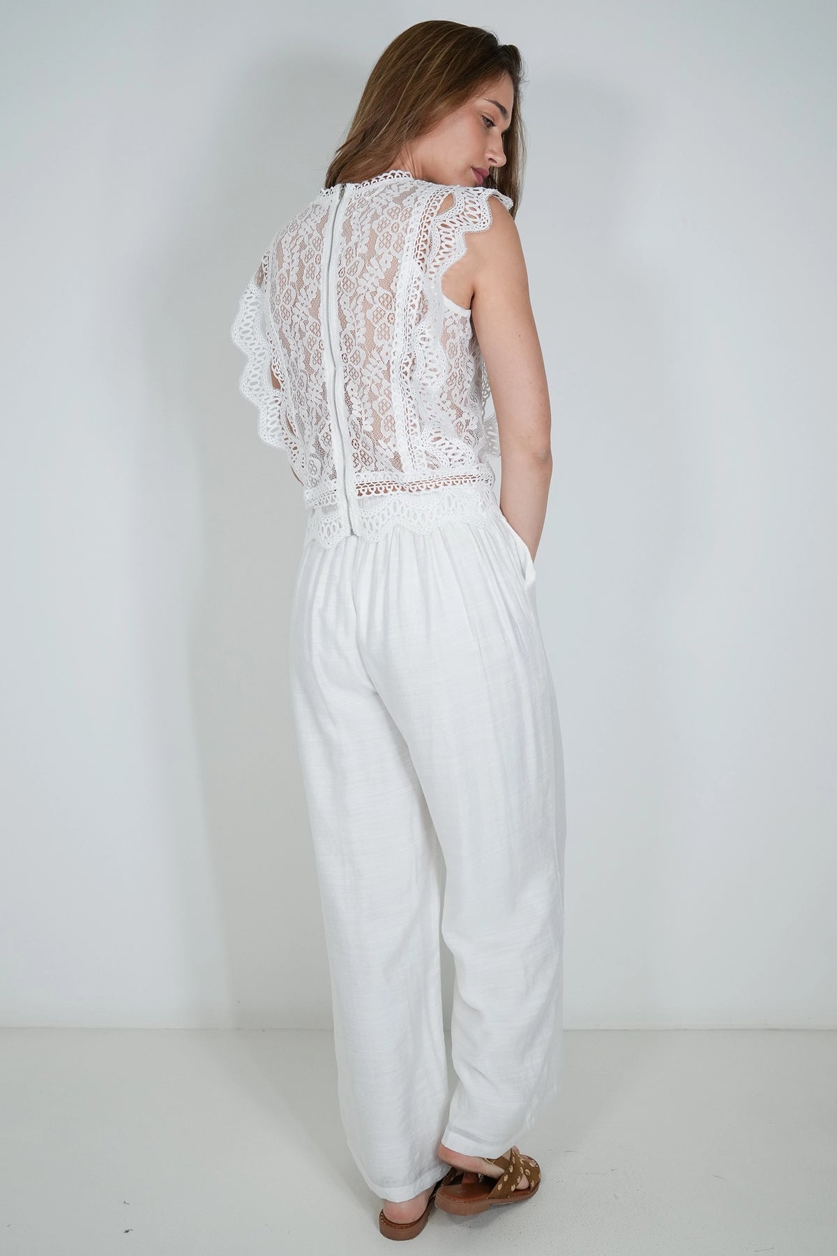 A woman wearing a white lace and linen jumpsuit, shown from the back against a plain white studio background.