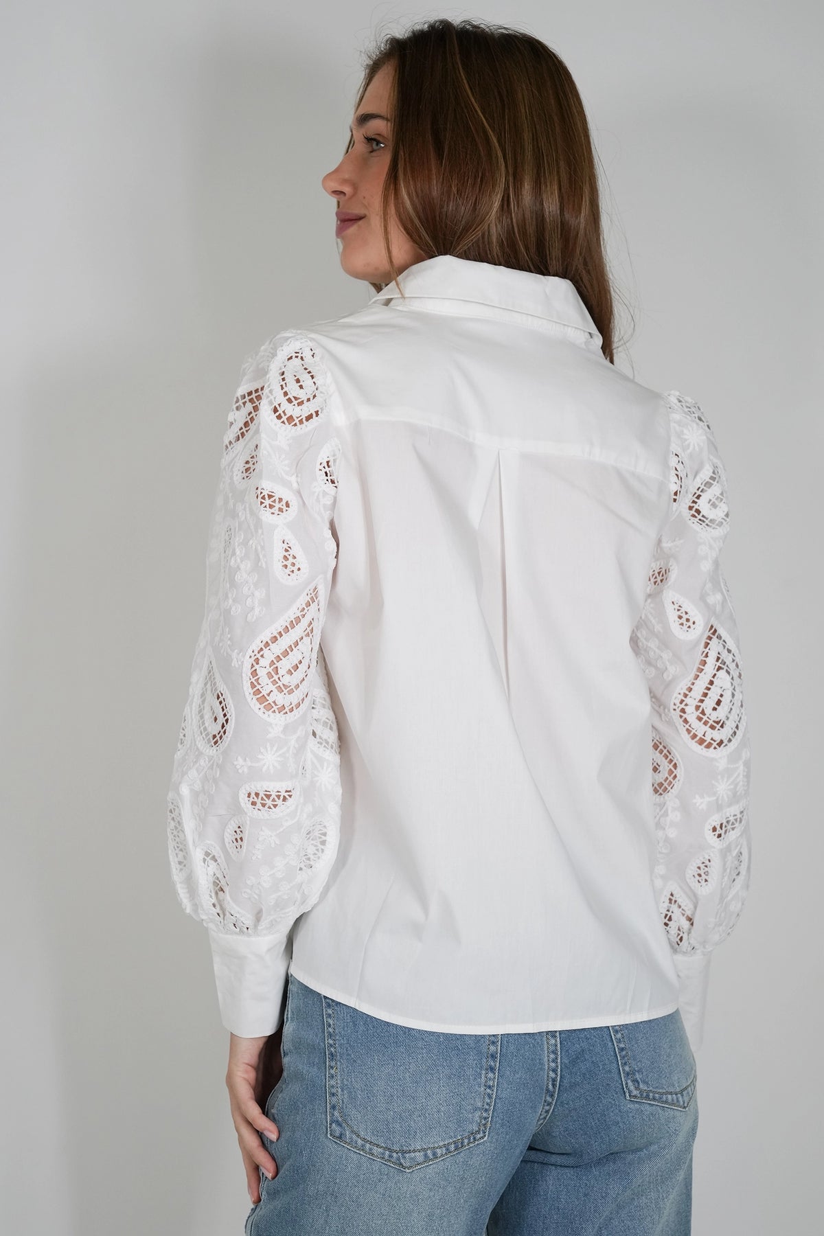 A woman shown from the back wearing a white long-sleeve blouse with intricate lace applique on the sleeves, paired with blue jeans, against a plain light gray studio background.