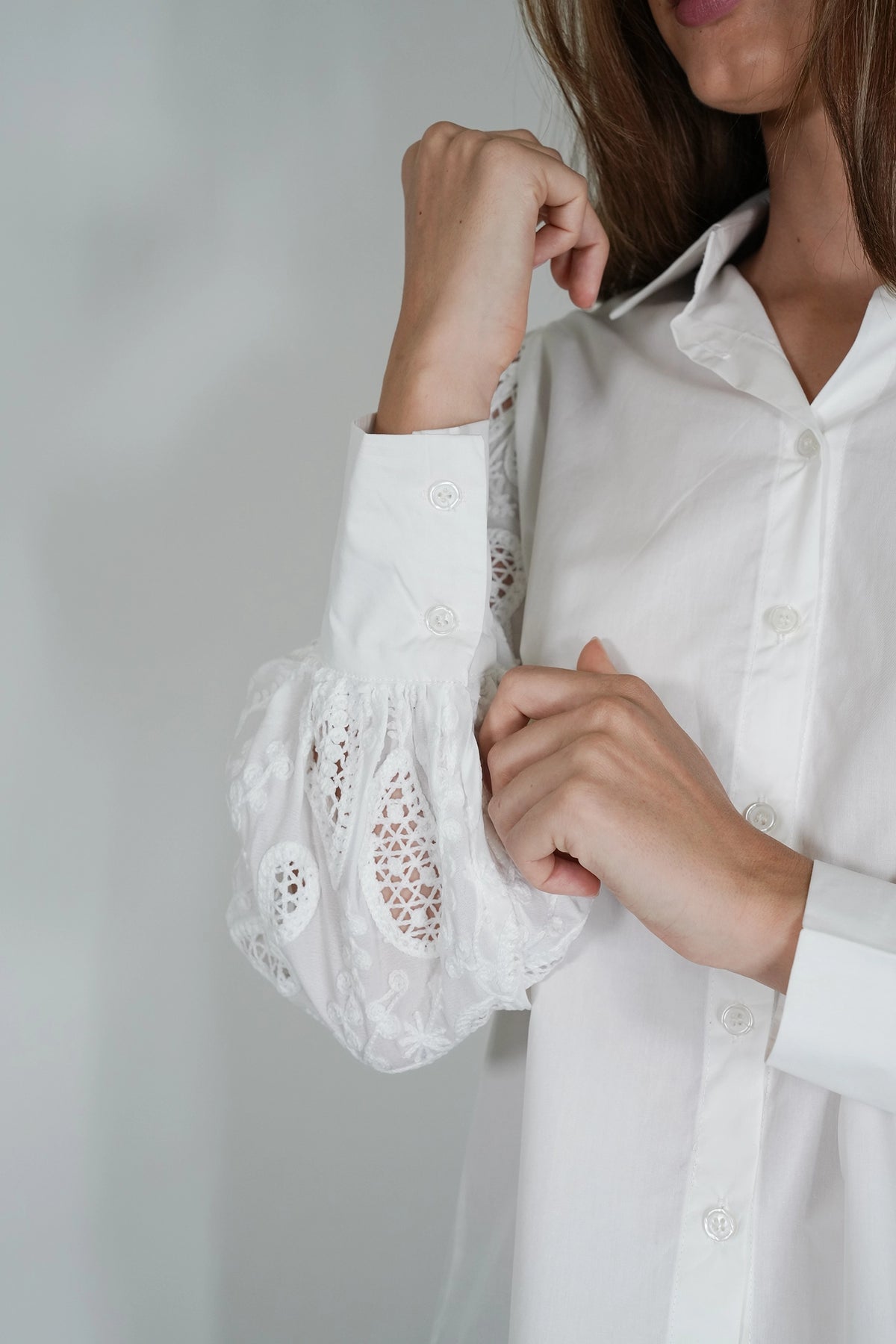 A close-up of a woman wearing a white long-sleeve button-up blouse with sheer lace puff sleeves, adjusting the sleeve with her hand against a light gray background.