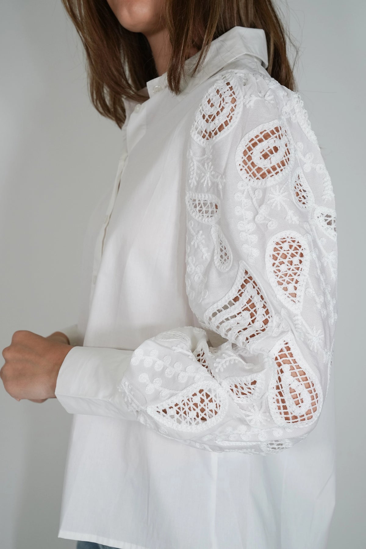 A close-up of a woman wearing a white long-sleeve blouse with intricate floral eyelet lace detailing on the sleeves, shown against a plain light gray background.