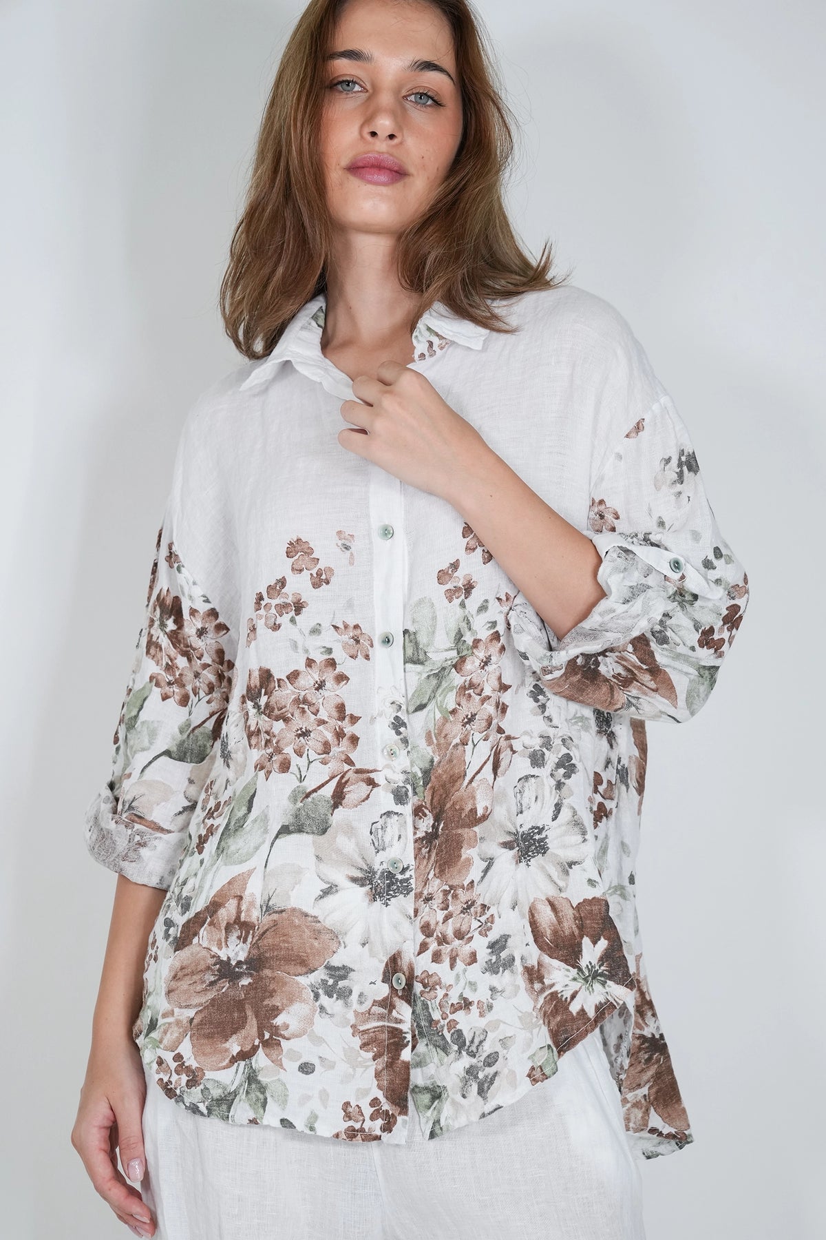 A woman wearing a white button-up blouse with a brown and green floral print, styled with white trousers against a light gray studio background.