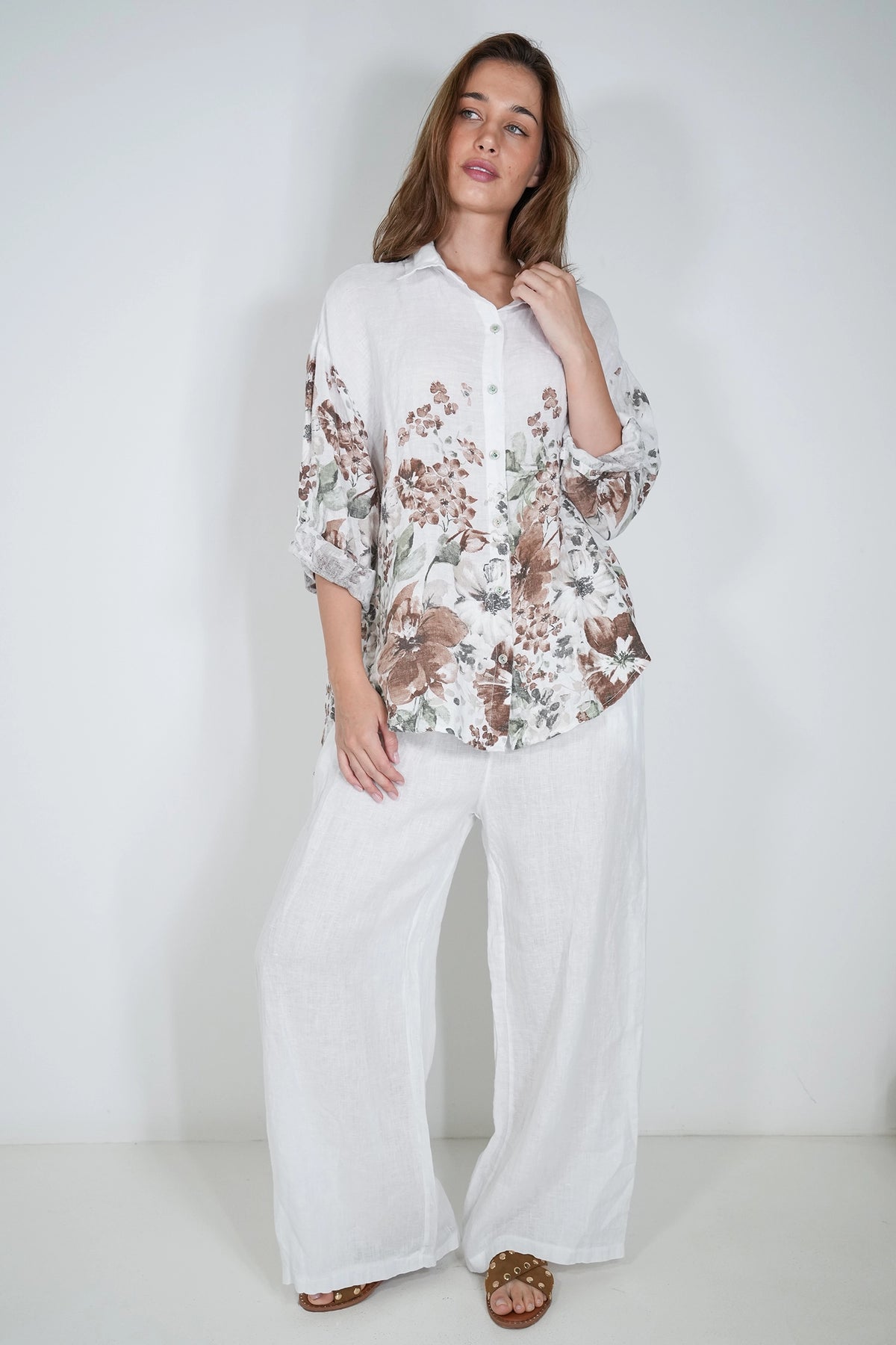 A woman modeling a white button-up shirt with a brown and beige floral print on the sleeves and hem, paired with white wide-leg pants and brown sandals, against a plain white studio background.