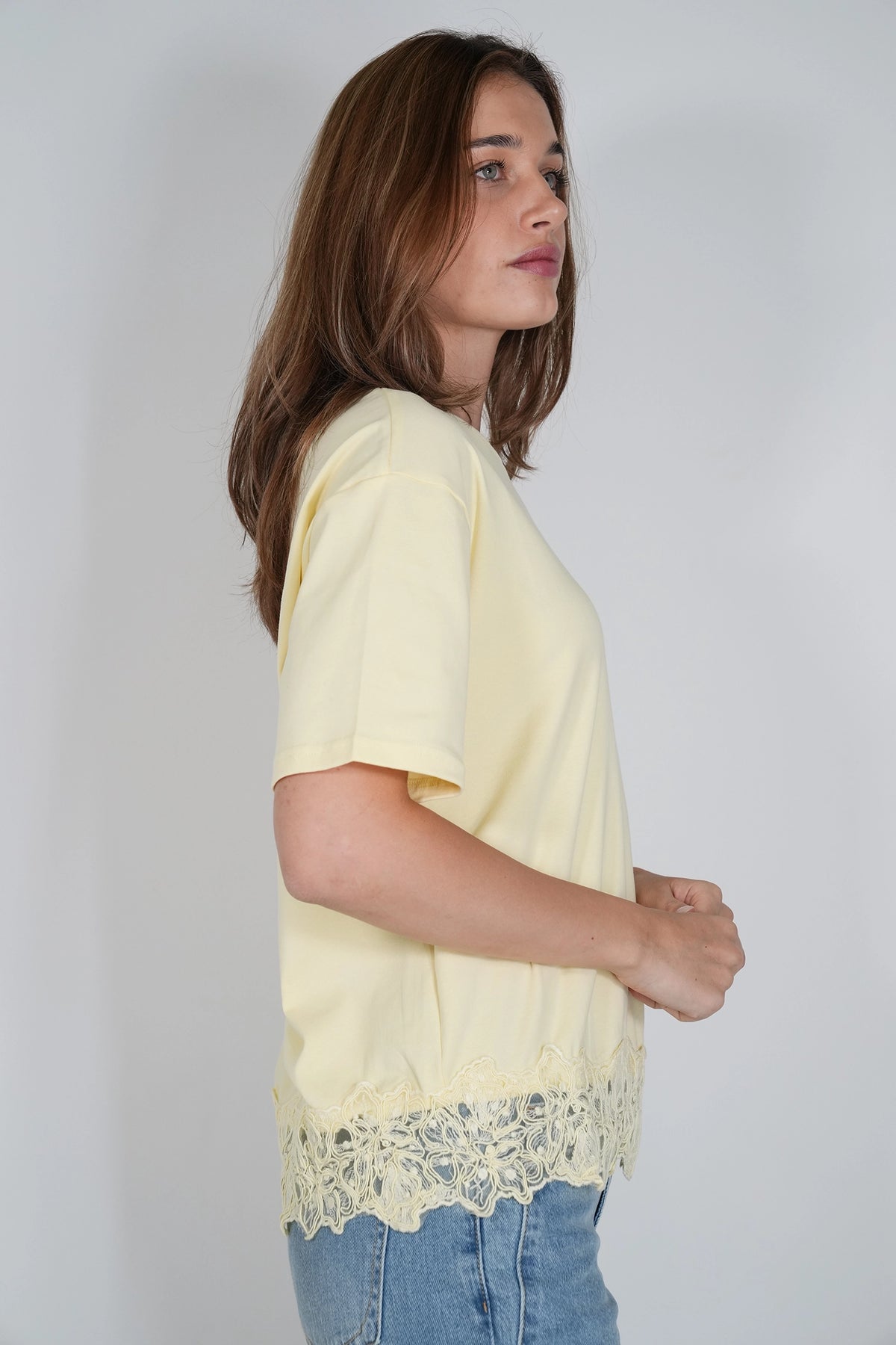 A woman wearing a pale yellow short-sleeve top with a scalloped lace hem, paired with blue jeans, shown in profile against a light gray studio background.