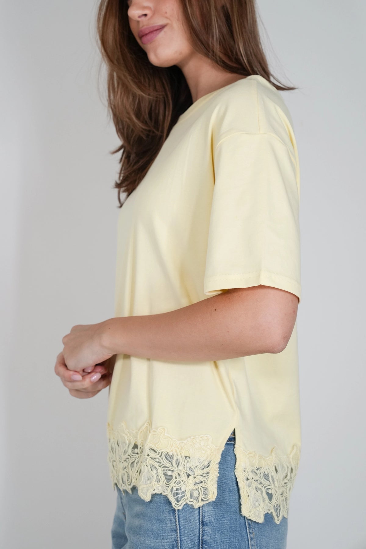 A woman wearing a pale yellow short-sleeve top with a scalloped lace hem, paired with blue jeans, shown from a side profile against a light gray background.