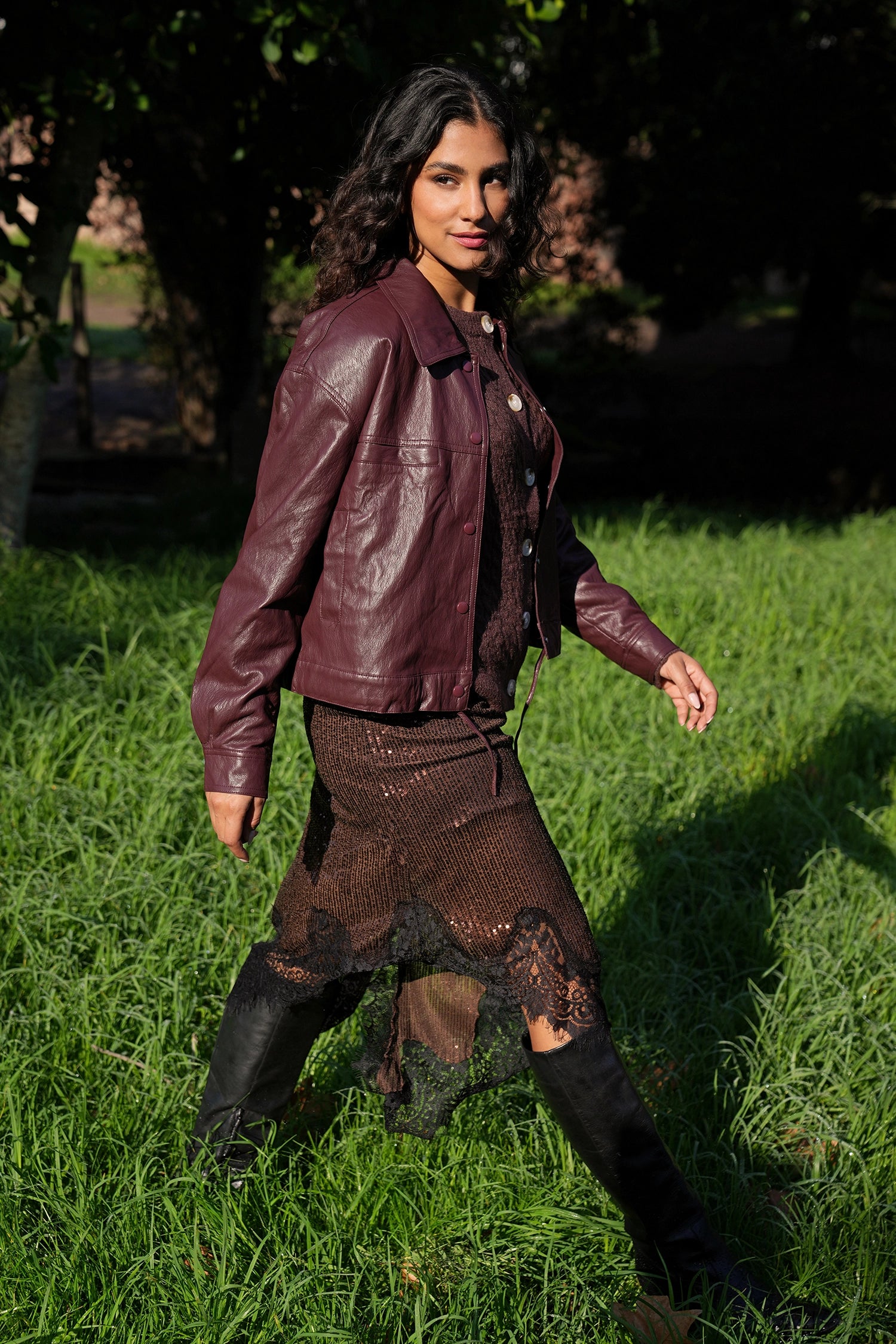 Woman in a burgundy leather jacket and black outfit standing in a grassy area.