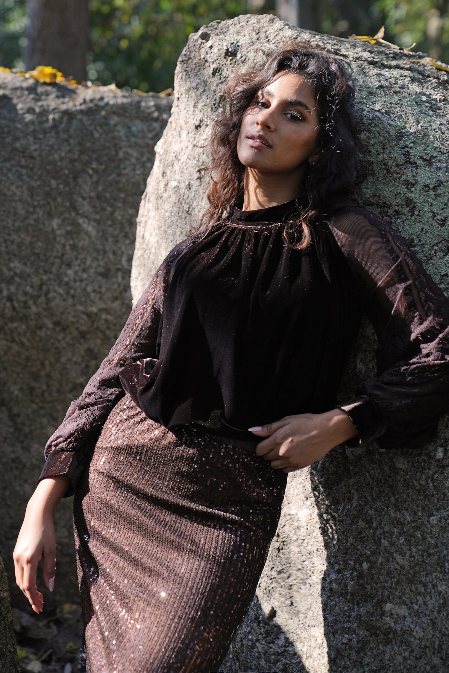 Person wearing a dark outfit with a brooch, standing against a stone wall.