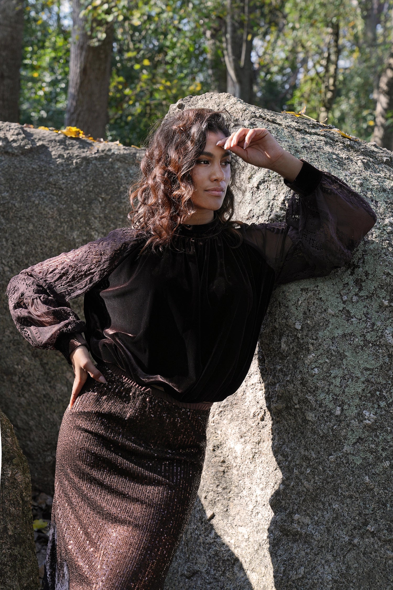 Woman in a dark outfit standing against a stone wall with trees in the background