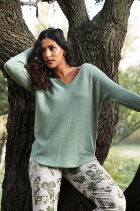 Woman in a light green sweater and patterned pants leaning against a tree in a natural setting