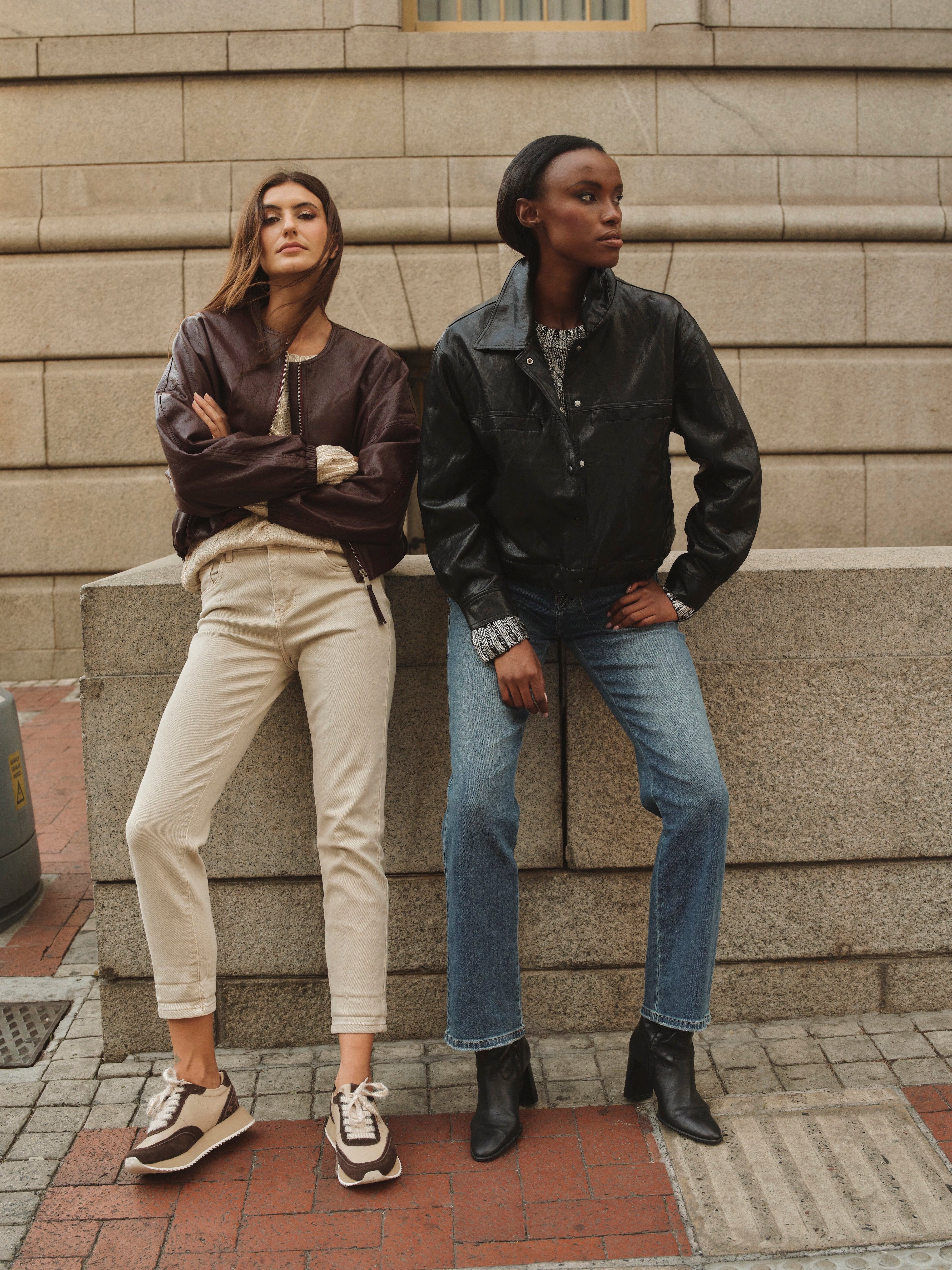 Two women standing against a stone wall, wearing jackets and jeans.