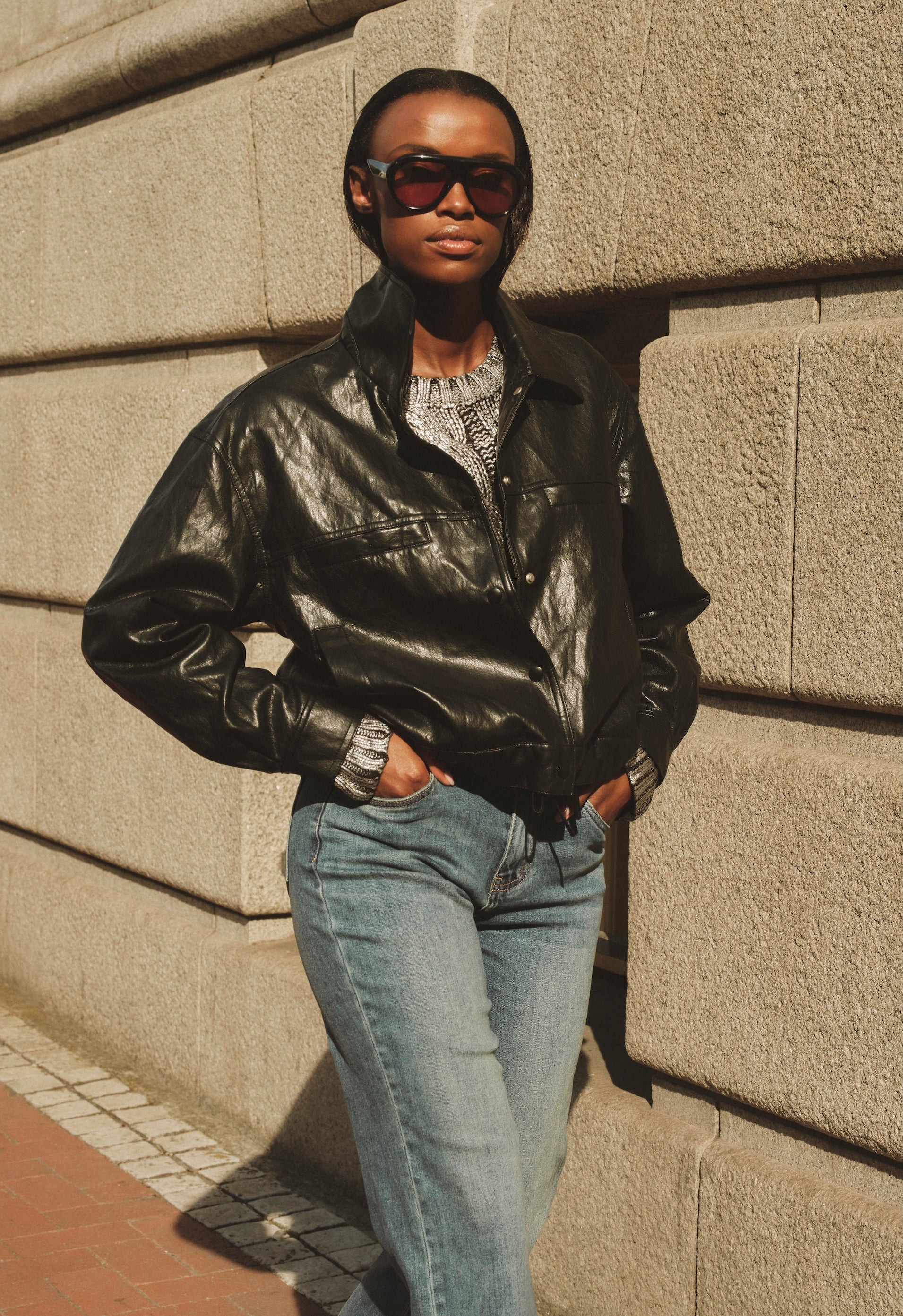 Person wearing a black leather jacket and sunglasses leaning against a textured wall.