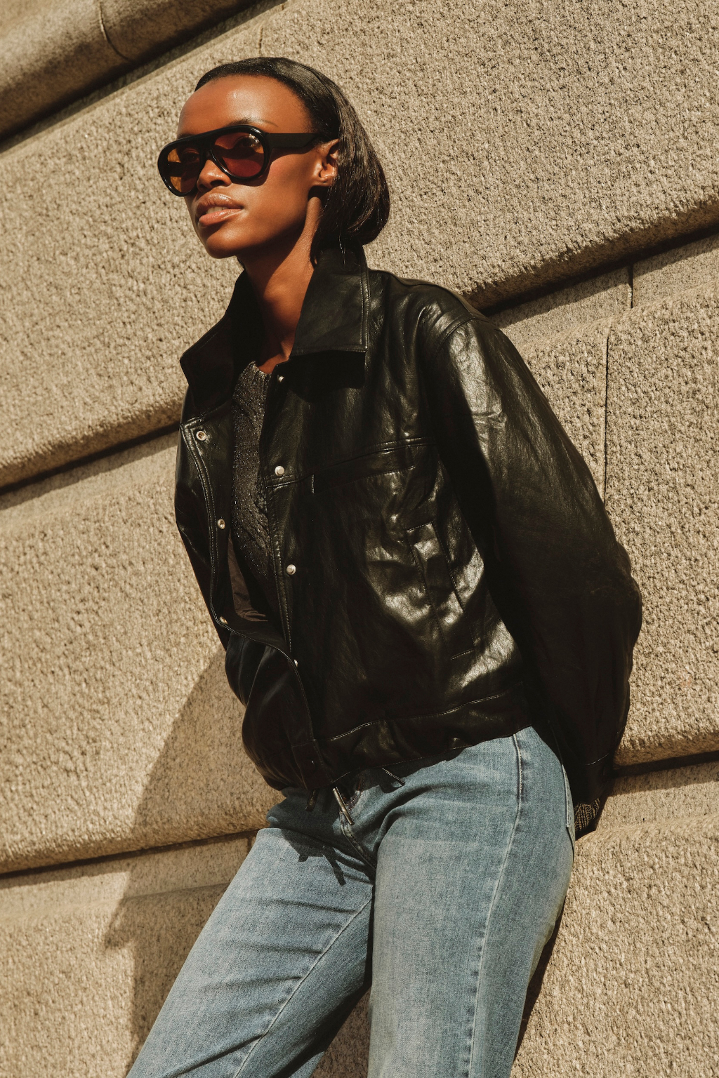 Person wearing a black leather jacket and blue jeans against a textured wall.