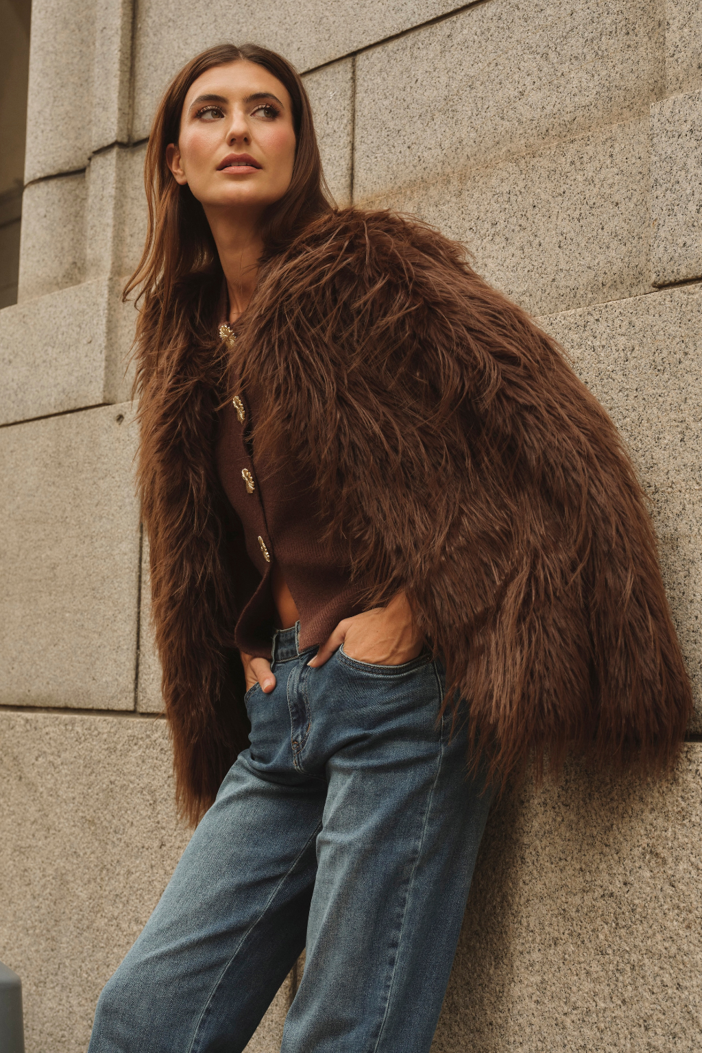 Woman wearing a brown fur coat and blue jeans against a stone wall.
