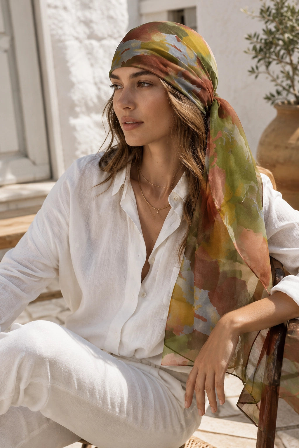 A woman wearing a white linen shirt and trousers sits outdoors, draped in a lightweight sheer scarf with an abstract watercolor pattern in green, yellow, orange, and brown tones.