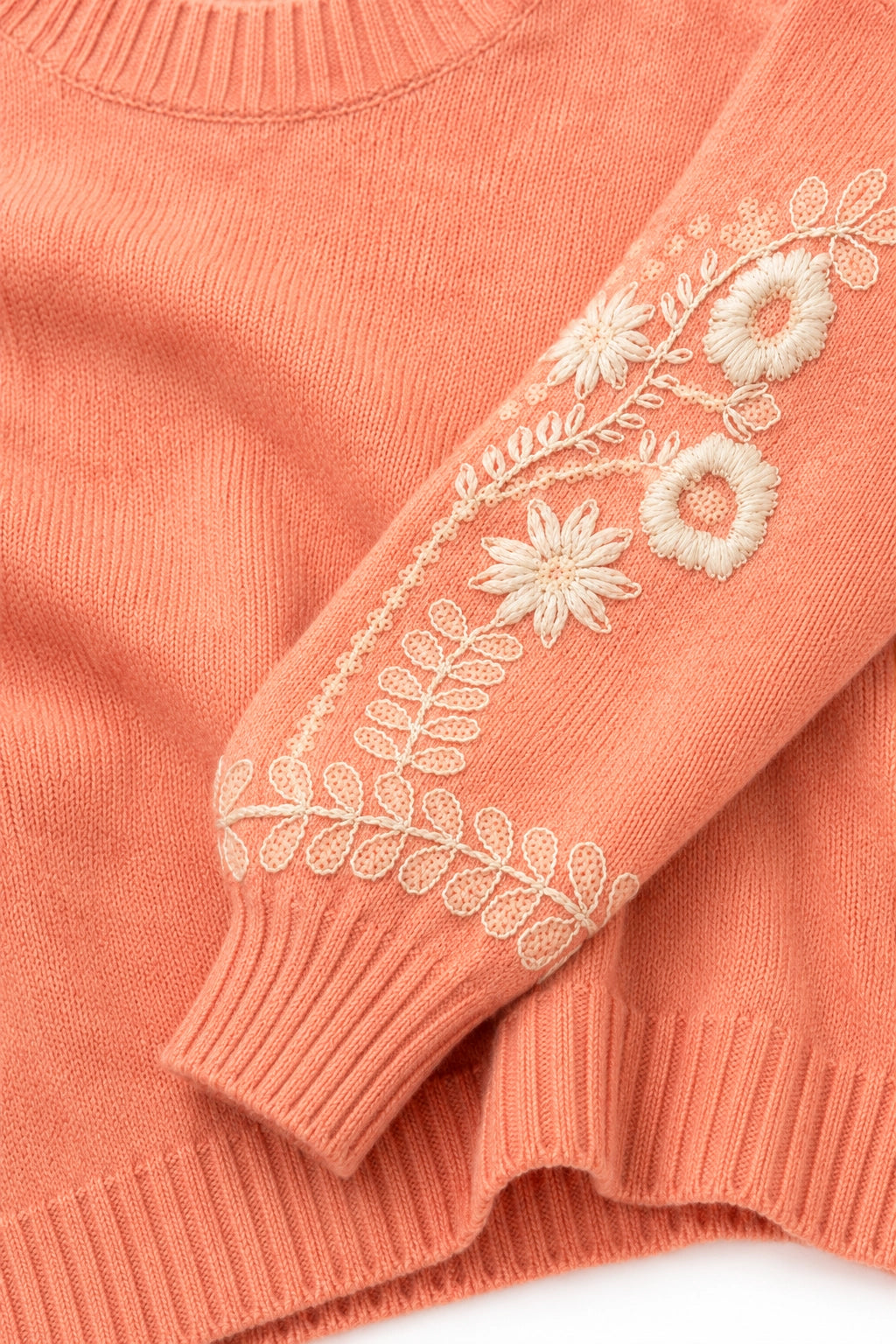 Close-up of a coral pink knit sweater with cream floral and leaf embroidery on the sleeve.